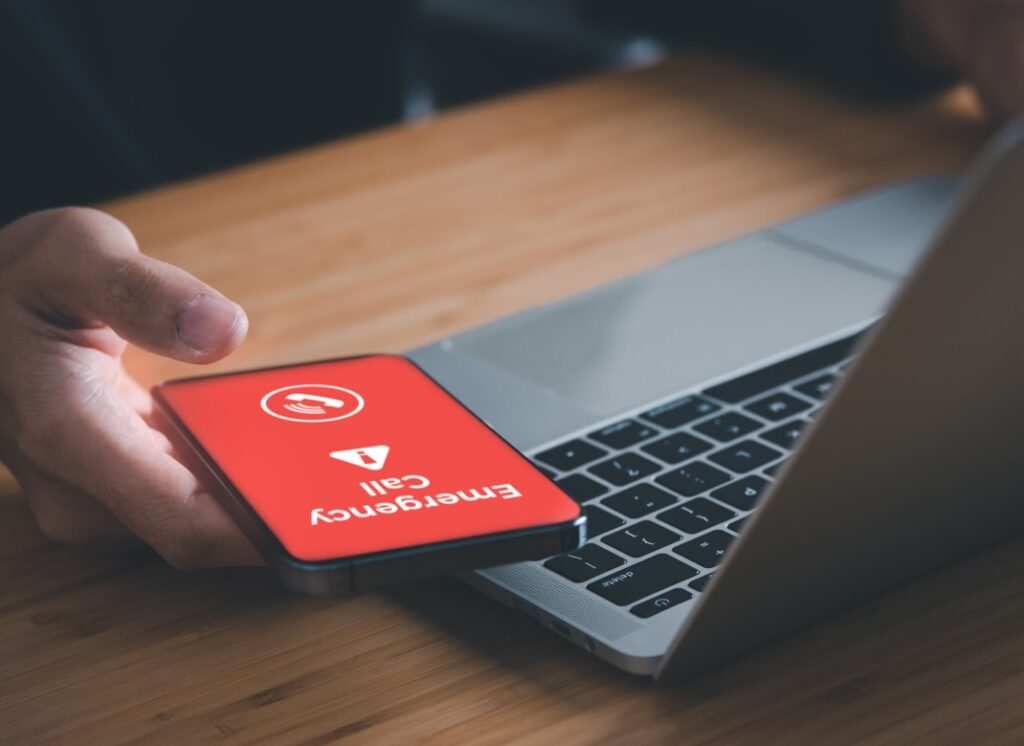 Hand holding a phone that says "emergency call" in front of a computer